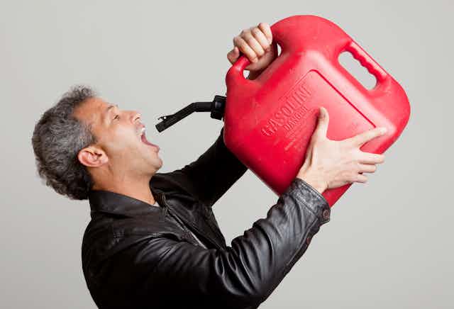 A white guy wearing a black jacket with his mouth open wide holds a red container of gasoline to his mouth