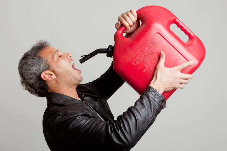 A white guy wearing a black jacket with his mouth open wide holds a red container of gasoline to his mouth