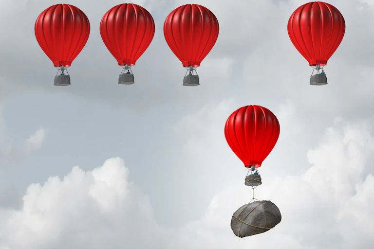 Five red hot-air balloons floating in a line, except for one which is dragged down by a large rock tied to it