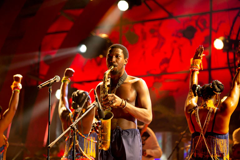 A red backdrop on a stage where a row of female dancers raise their fists in the air, in front of them a bare chested man plays saxophone into a microphone.
