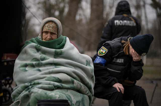 An elderly woman sits under a green blanket as a police officer cries into her hands beside her.