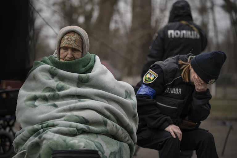 An elderly woman sits under a green blanket as a police officer cries into her hands beside her.