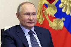 A balding man smiles with a red, blue and gold flag behind him.