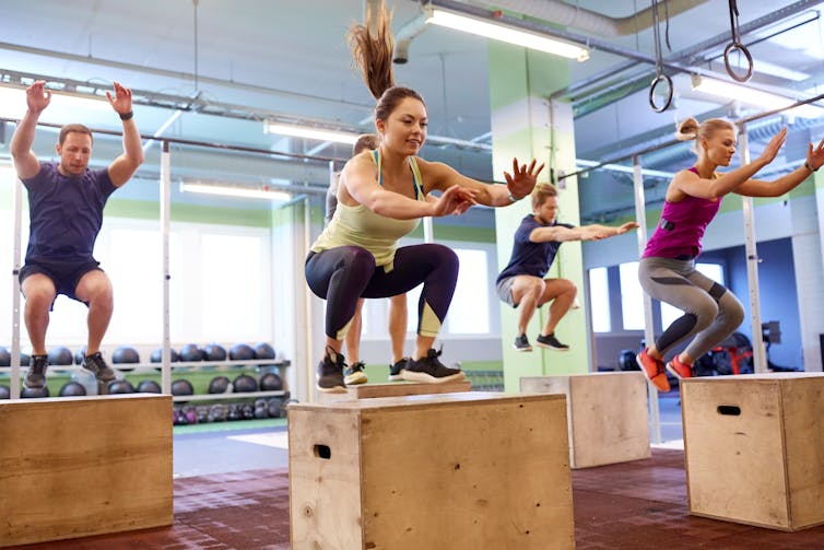 A group of athletic young people perform box jumps in a gym.