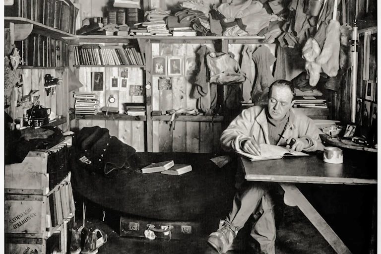 Explorere sits at desk in his book filled room.