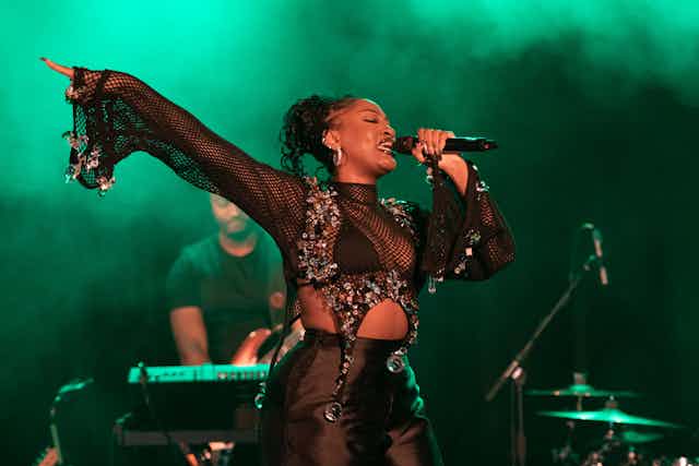 A woman dressed in a black two-piece with exposed midriff sings into a microphone, her arm outstretched and a finger pointed, behind her a man playing keyboard and green lights with smoke.