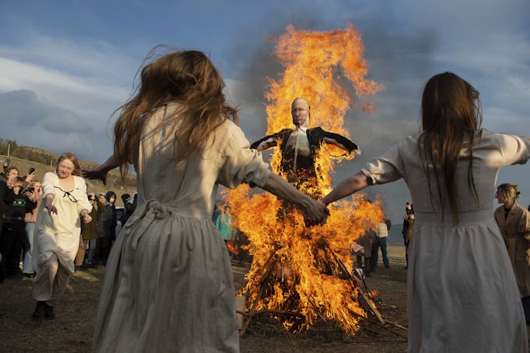 Two long-haired girls join hands and dance in front of a burning effigy of Vladimir Putin.