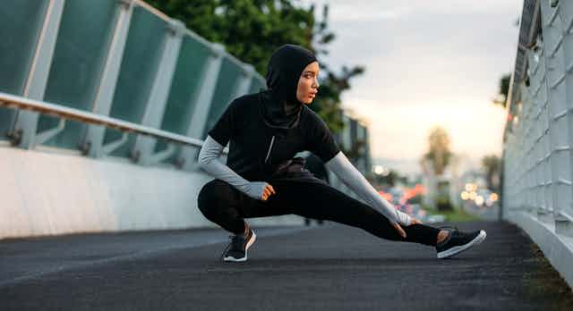 A girl in a black hijab and running closes does stretches on a bridge.