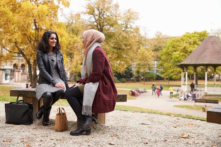 A woman in a leather jacket sits on a bench in a park with another woman in a pink hijab and marroon coat.