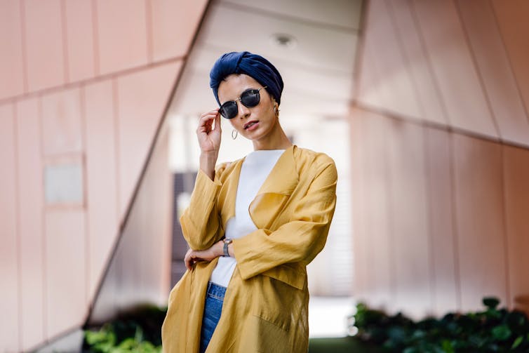 A woman in a blue headscarf and yellow coat poses in front of a pink building.