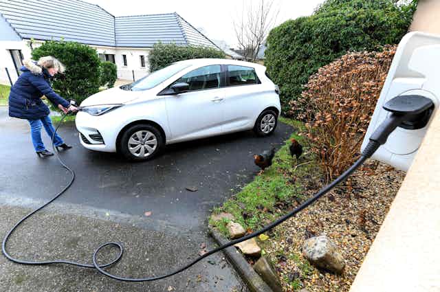 A woman charges an electric car in her driveway from a cord plugged into an exterior socket on her house