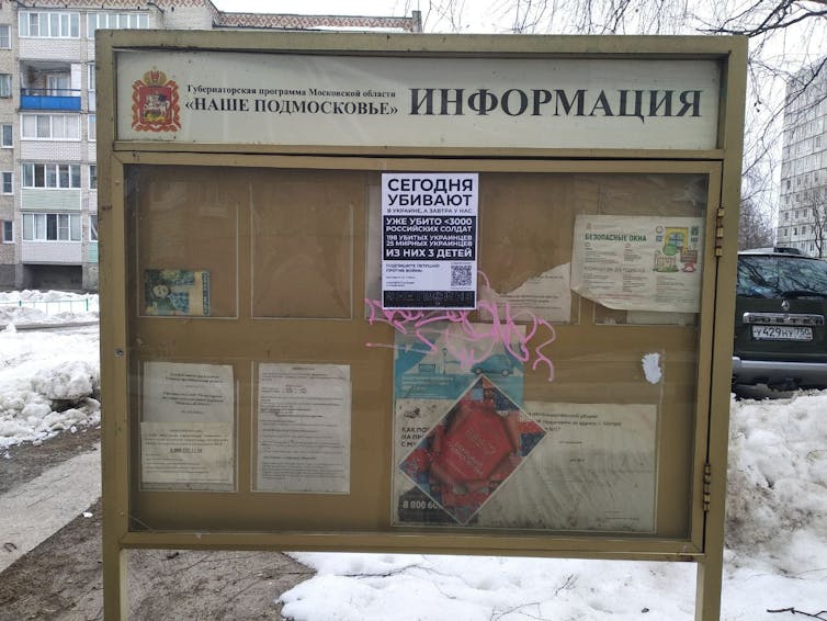 A glass case community billboard stand is seen outdoors with a paper sigh with Russian writing glued onto it.