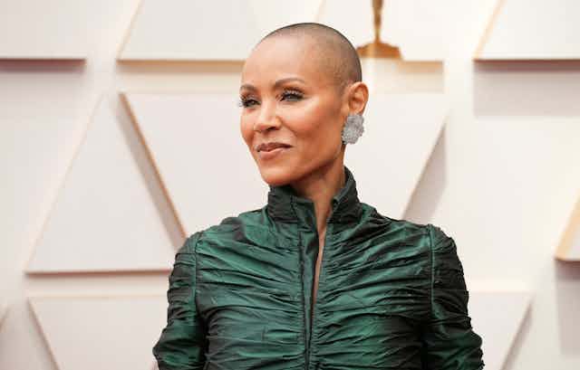 Jada Pinkett Smith with a shaved head and wearing a green ballgown poses on the red carpet.