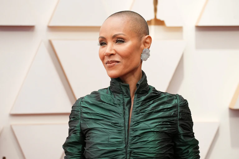 Jada Pinkett Smith with a shaved head and wearing a green ballgown poses on the red carpet.