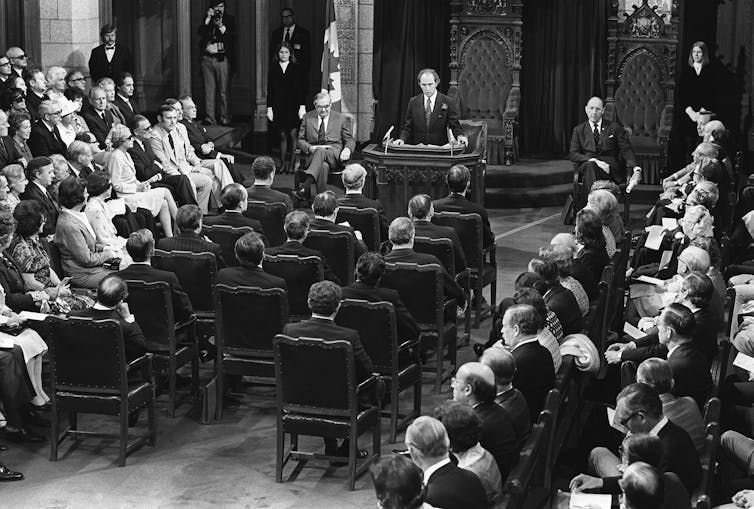 Men and some women sit in a parliamentary chamber in this black and white photo with one man behind a podium.