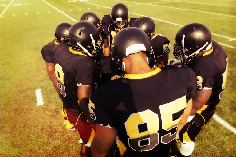 A huddle of football players in black uniforms and helmets on the field.