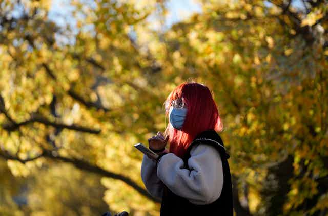 A woman with hair dyed bright red wears a mask as she uses her phone