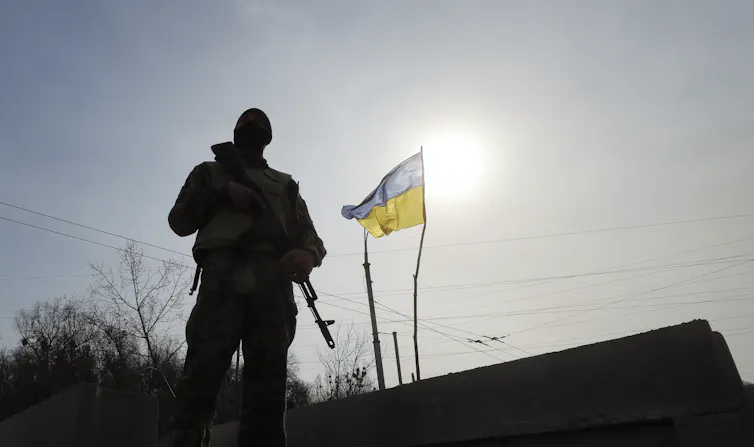 A Ukrainian soldier in silhouette against the setting sun with a national flag in the background.