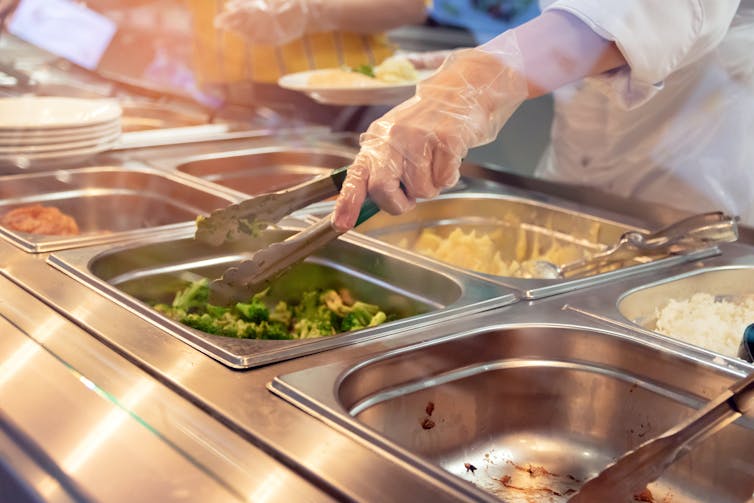 A gloved hand reaches for broccoli at a cafeteria serving hatch.