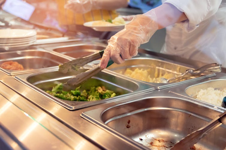 A gloved hand reaches for broccoli at a cafeteria serving hatch.