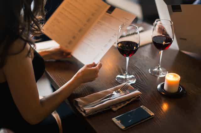 A woman reads a menu at a table with a glass of wine.