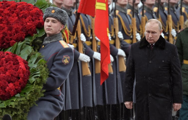 A man in a dark cloth coat next to a line of soldiers, several of whom are carrying wreaths.