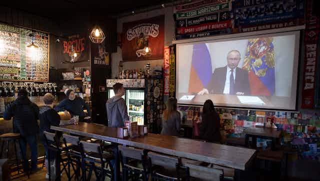 A balding man appears on a TV screen as people in a bar watch.