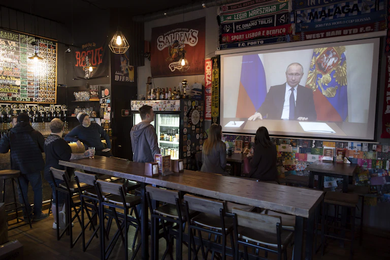 A balding man appears on a TV screen as people in a bar watch.