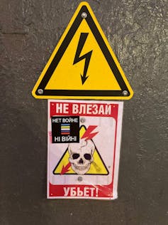 A skull with a bolt going through it is seen affixed to a grey surface under a triangular public safety sign.