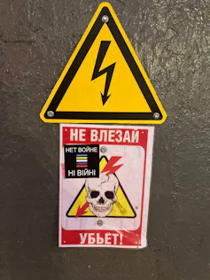 A skull with a bolt going through it is seen affixed to a grey surface under a triangular public safety sign.
