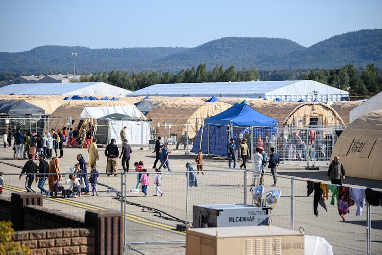 Children and adults are seen from a distance in front of beige, white and blue tents, all fenced in