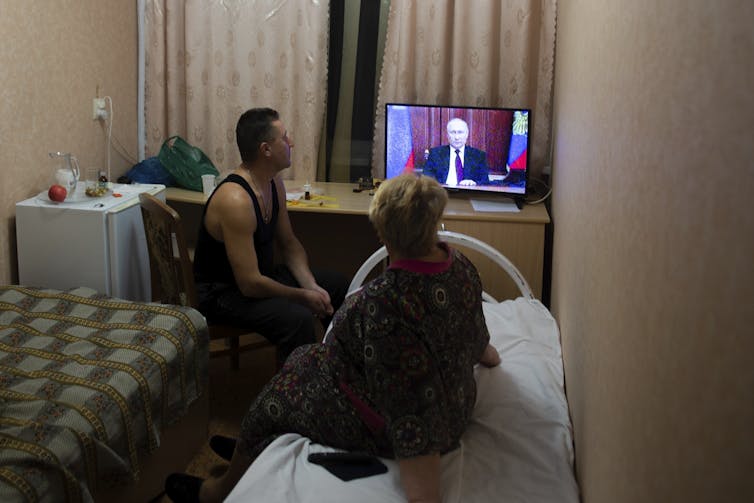 A man and woman sit in a dark room watching a balding man on a television set.