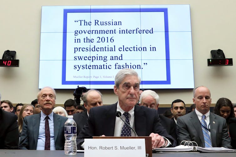 A man sits in front of a microphone with a screen behind him that says Russia interfered in the U.S. presidential election.