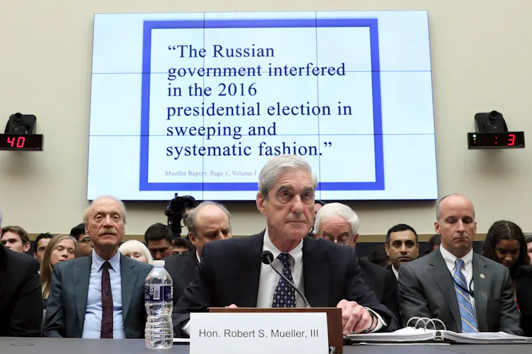 A man sits in front of a microphone with a screen behind him that says Russia interfered in the U.S. presidential election.