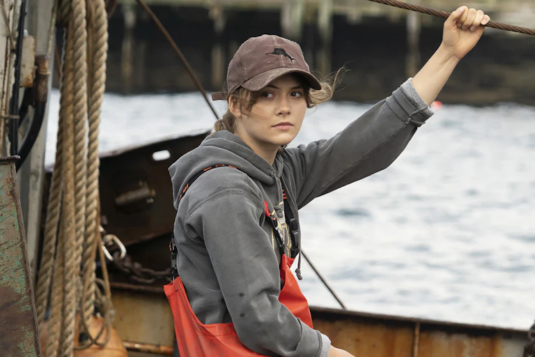 A young woman in a cap on a boat holds a rope line.