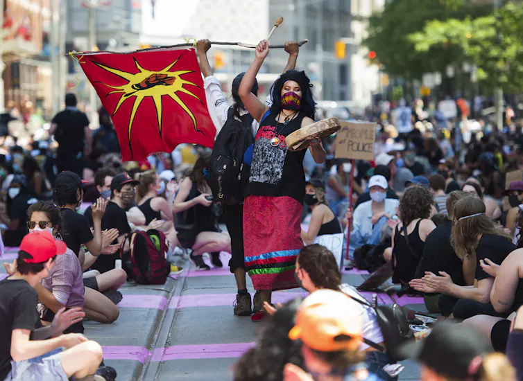 A woman stands amongst people sitting, wearing a ribbon skirt as she drums.