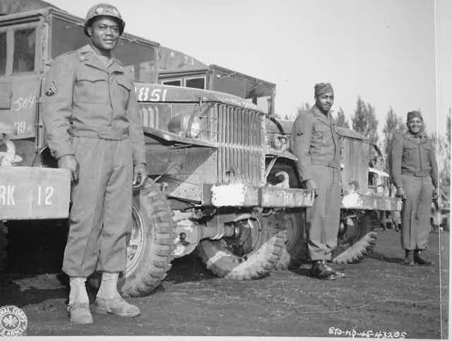 Three black soldiers are standing in front of several military trucks.t 