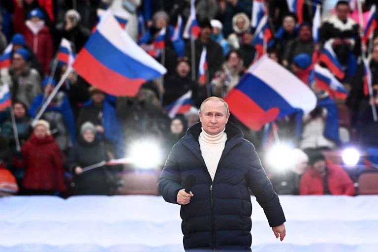 A man in a dark winter coat and white turtleneck standing on stage outside before a flag-waving crowd.
