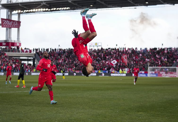 A man in a Team Canada uniform does a backflip with a full crowd in the background.