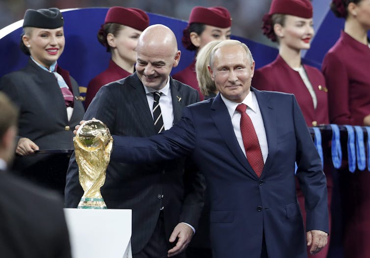 Putin reaches to the right and grabs the World Cup trophy.