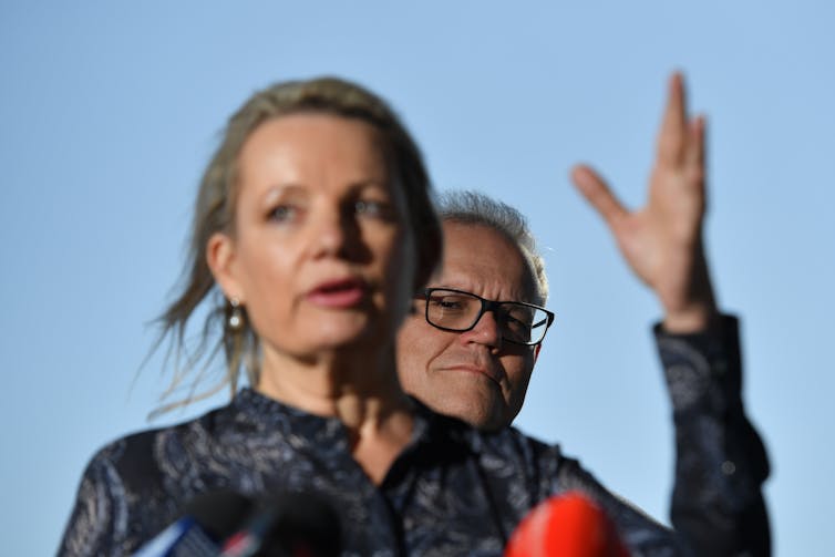 man in glasses stands behind woman with hand in air