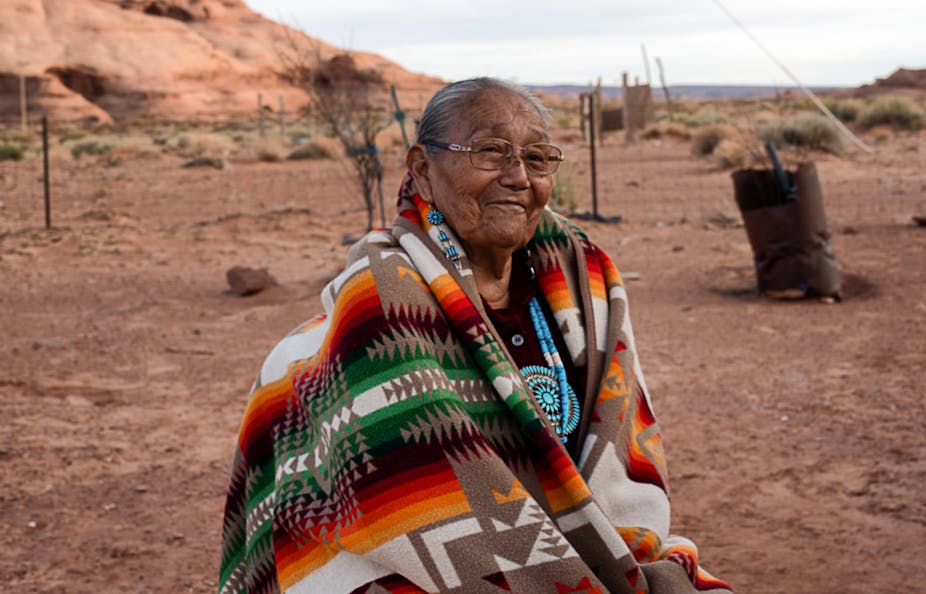 Smiling 85-year-old Native American Navajo grandmother posing for a portrait in a yard surrounded by red rocks in Monument Valley, Arizona.
