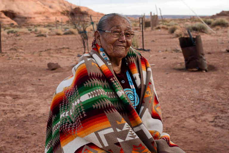Smiling 85-year-old Native American Navajo grandmother posing for a portrait in a yard surrounded by red rocks in Monument Valley, Arizona.