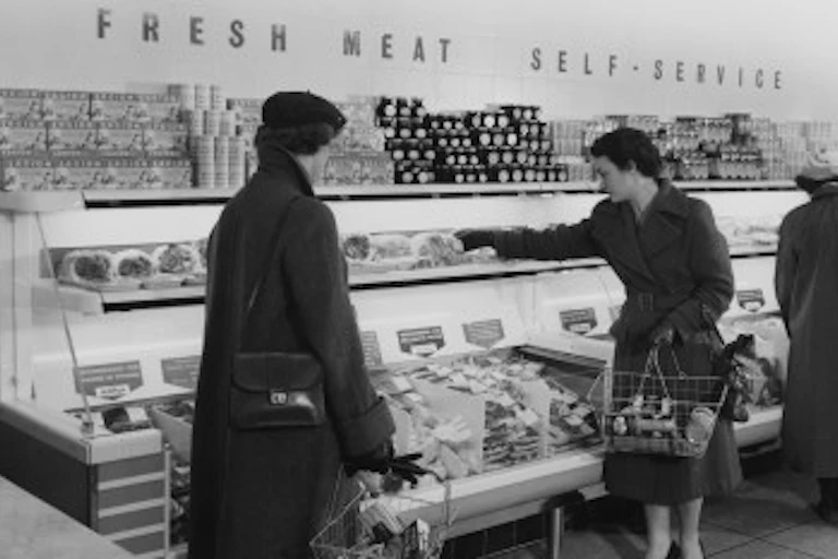 Women holding shopping baskets in front of a counter with a sign saying fresh meat self-service.