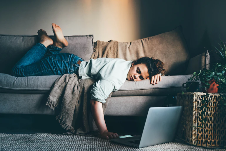 A woman who looks bored and tired lying on a sofa looking at her laptop on the floor below her.