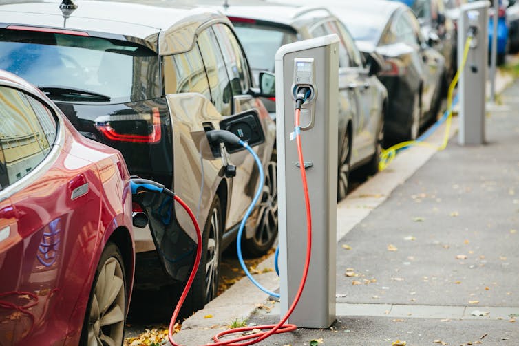 A row of cars parked along a street, with several plugged into small charging towers.