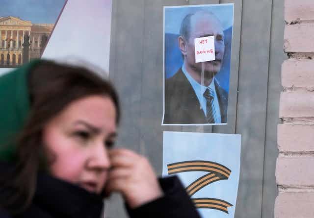 A dark-haired woman walks past a window with a photo of a man that has a sticker on his face saying 'no war' in russian and a Z under the photo that represents support for russia's military