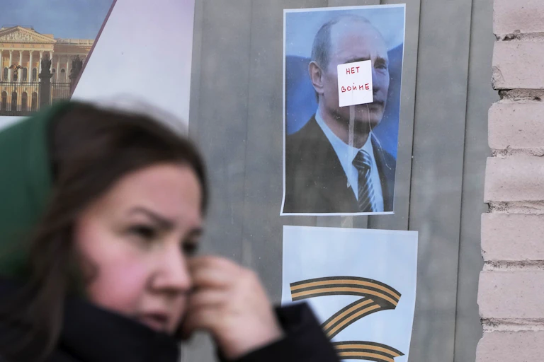 A dark-haired woman walks past a window with a photo of a man that has a sticker on his face saying 'no war' in russian and a Z under the photo that represents support for russia's military