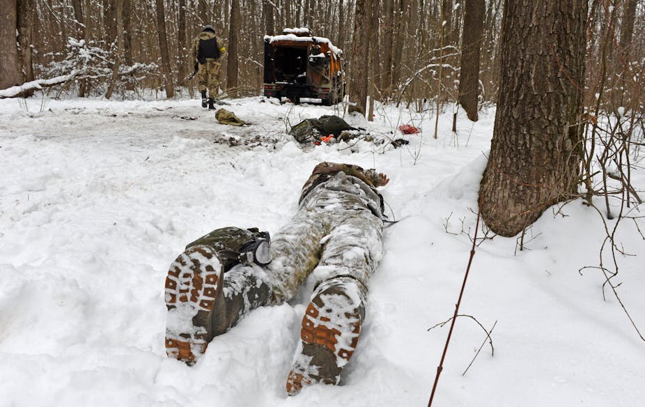 A snow-covered body in boots and fatigues lies next to a tree with a destroyed Russian vehicle in the background.