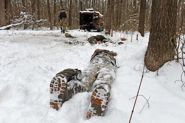 A snow-covered body in boots and fatigues lies next to a tree with a destroyed Russian vehicle in the background.